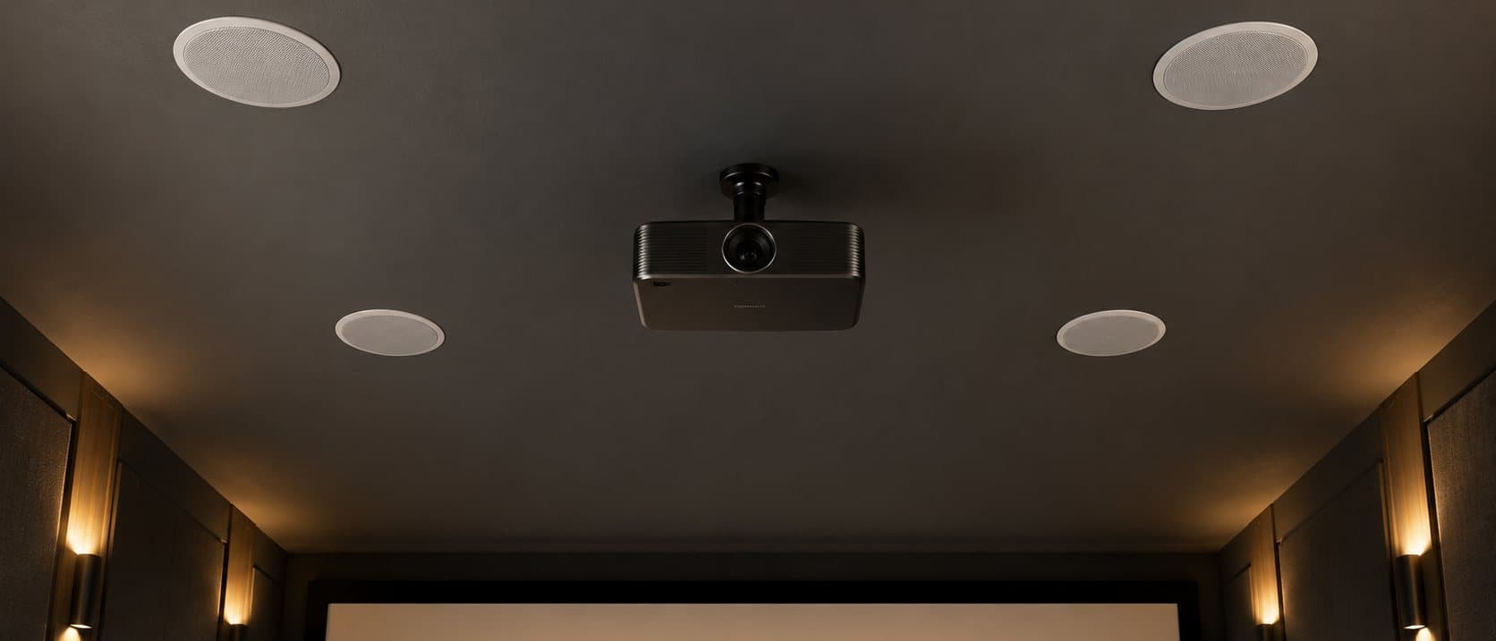 Looking up at a dark theater ceiling with four round in-ceiling speakers around a mounted projector, sconces glowing below